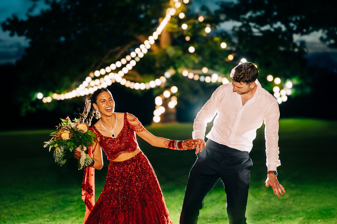 Bride in red lehenga and groom dancing together on the lawn under festoon lights at Rathsallagh House at night