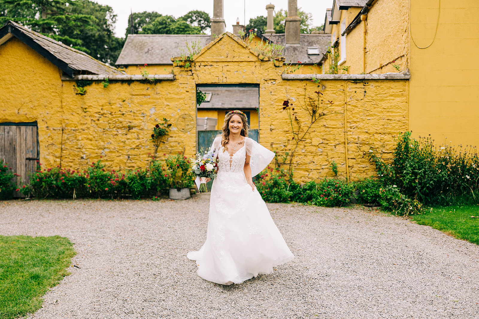 Magdalena & Ronan | Martinstown House bride walking in front of the yellow walled garden at Martinstown House Kildare holding a wildflower bouquet and wearing a flower crown